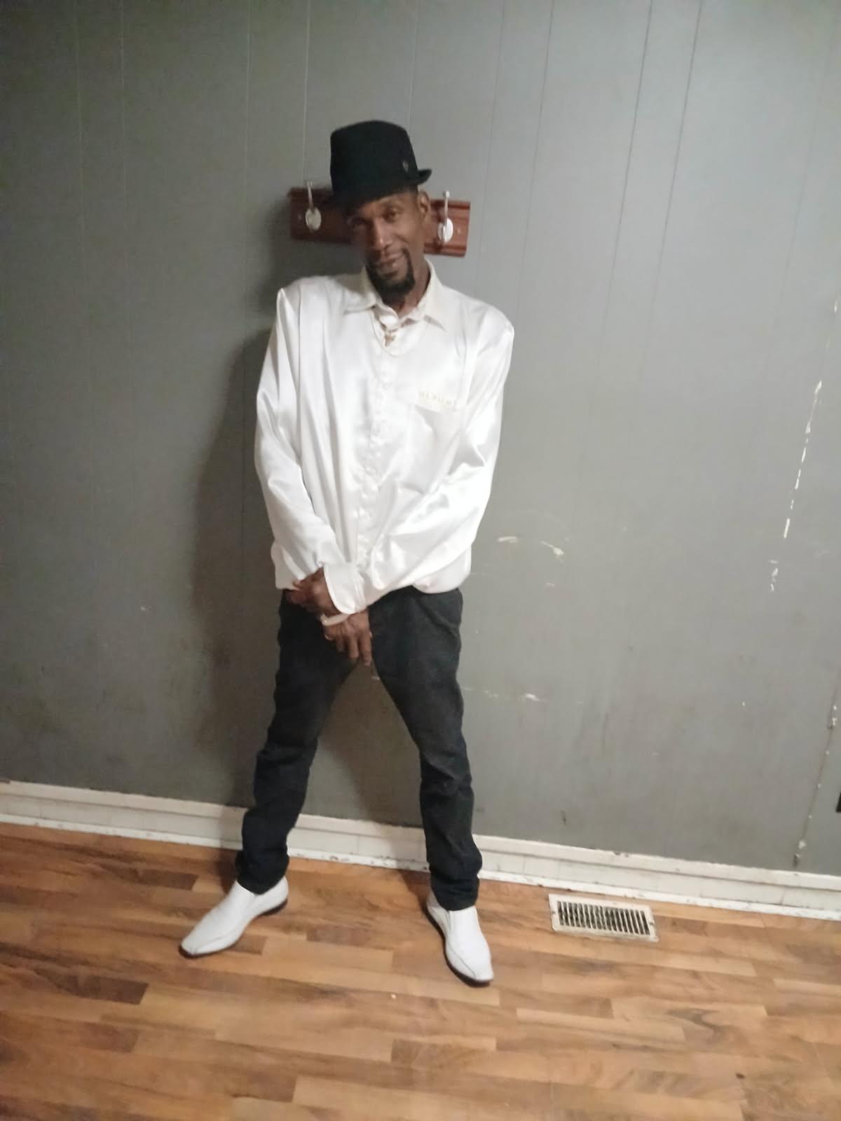 Introducing The General Da Jamaican Boy, a rising star in the music scene, captured in a striking image that embodies his unique style and charisma. Dressed in a crisp white shirt and a classic black hat, he stands confidently against a textured wall, symbolizing the grit and determination behind his artistry.